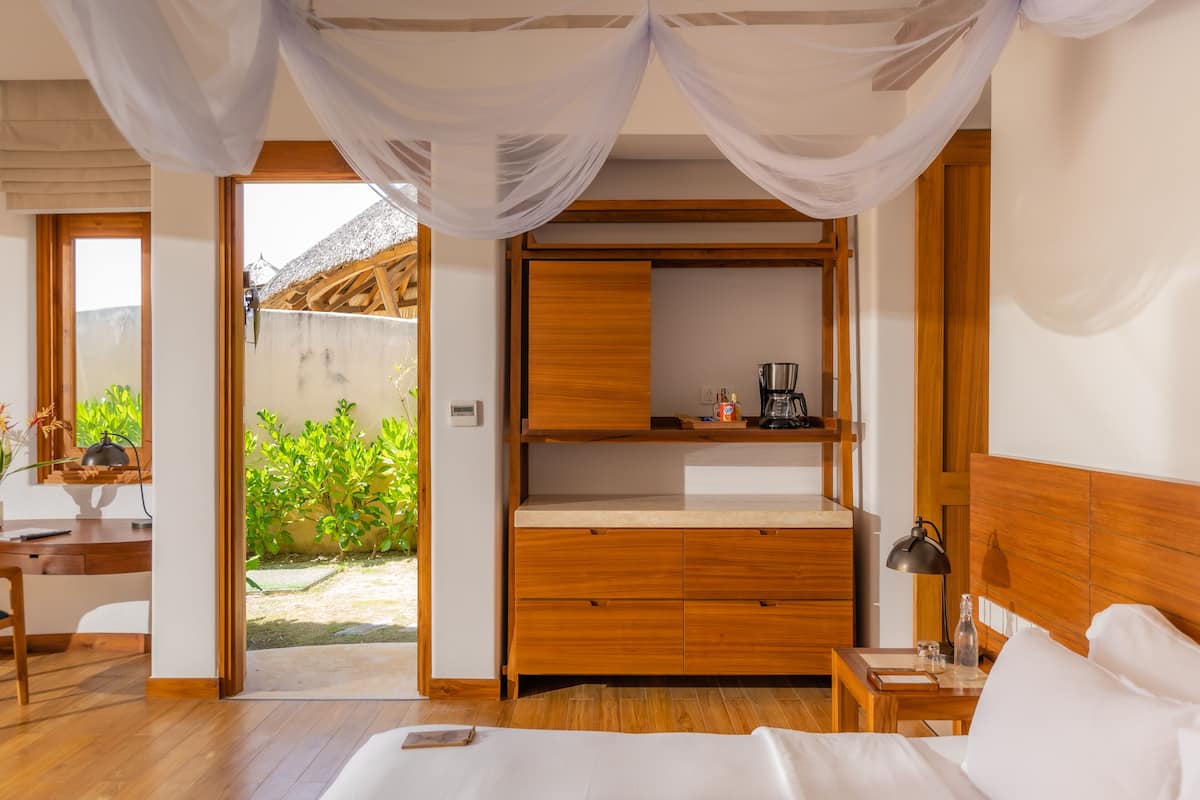 A bedroom with a wooden headboard, a bed, a nightstand, and a desk. There is a window with a view of greenery outside.