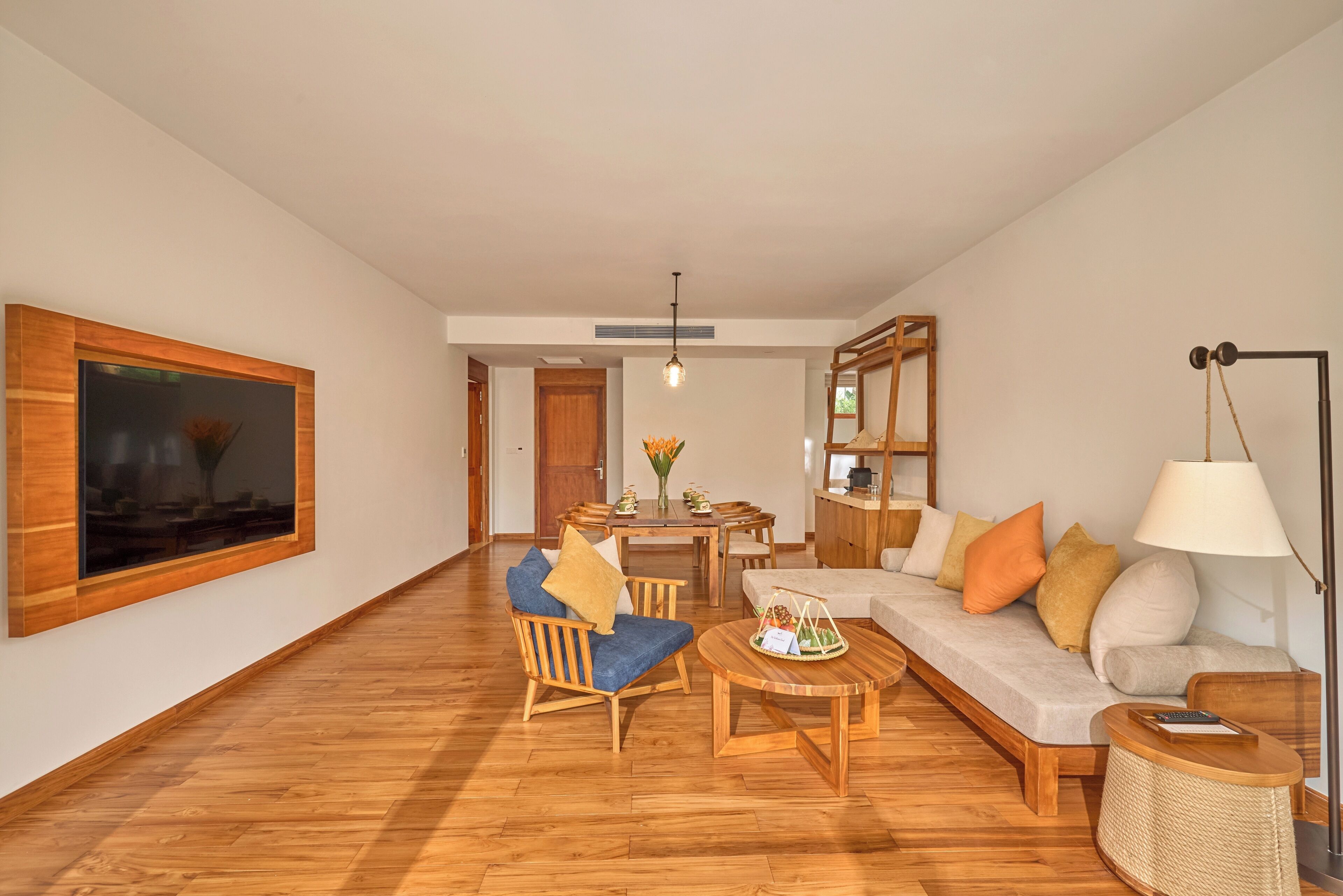 A spacious living room with a wooden floor, a large TV mounted on the wall, a dining area with a table and chairs, and a sofa with cushions.
