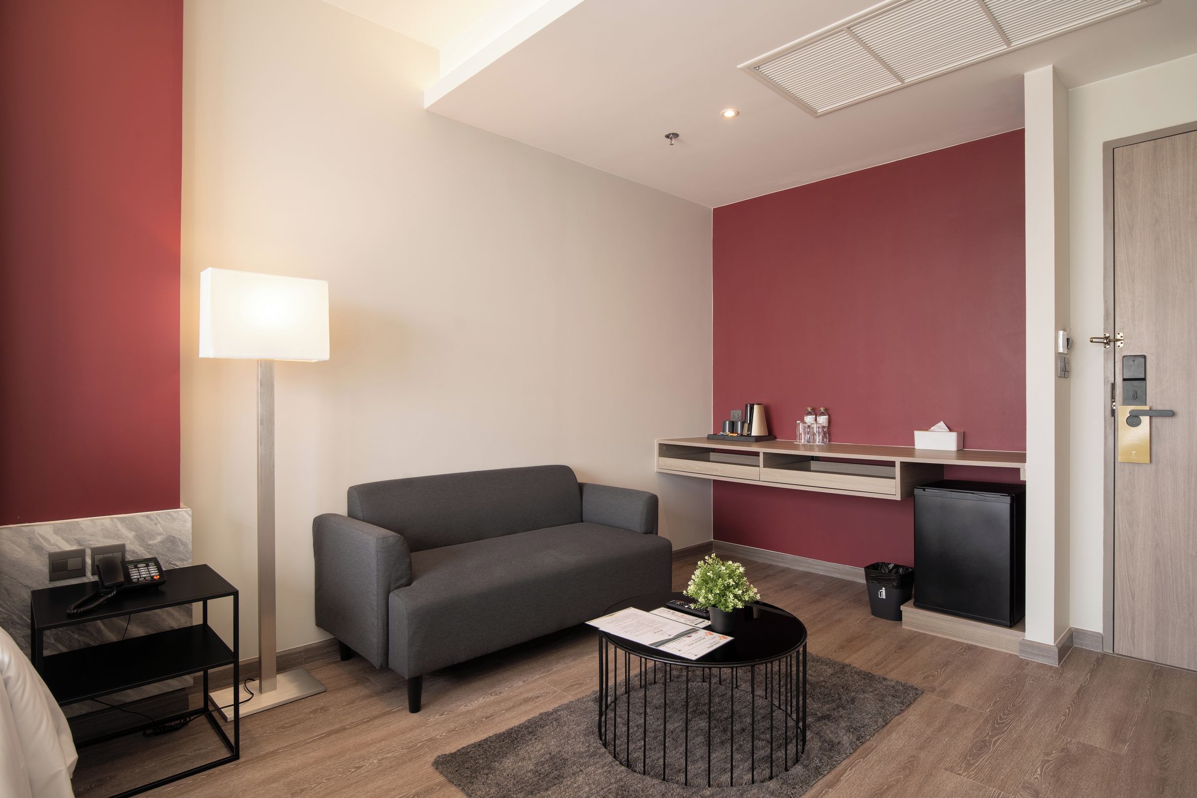 A modern living room with a grey sofa, a small round table with a plant, a standing lamp, and a kitchenette area with a fridge and a wooden cabinet.