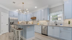 A modern kitchen with light blue cabinets, stainless steel appliances, and a central island with bar stools. - Beautiful new home just mins to the beach, aquarium, BUC-EES, and Downtown! (Gulfport)