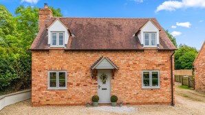 A two-story brick house with a gabled roof, a front door with a small porch, and two windows on the upper floor. - Orchard Cottage, Alderton - sleeps 6 guests  in 3 bedrooms (Alderton)