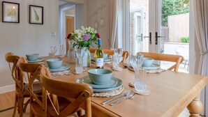 A dining table set with plates, bowls, cutlery, and a bottle of wine, surrounded by wooden chairs. - Orchard Cottage, Alderton - sleeps 6 guests  in 3 bedrooms (Alderton)