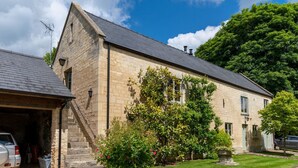 A stone building with a steep roof, a garage, and a well-maintained garden with flowering plants. - Stable Cottage at Freams Farm, Painswick - sleeps 5 guests  in 3 bedrooms (Painswick)