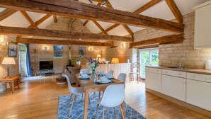 A rustic interior with exposed wooden beams, a stone fireplace, a dining area with white chairs, and a kitchen with white cabinets. - Stable Cottage at Freams Farm, Painswick - sleeps 5 guests  in 3 bedrooms (Painswick)