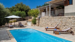 A swimming pool with lounge chairs and umbrellas, a stone wall, and a building with a balcony. - Lovely Sicilian-style villa with pool and sea view, 3 Km from the beach  (Scicli)