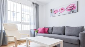 A modern living room with a grey sofa, a white coffee table, and a white armchair. There is a window with grey curtains, and a painting of pink flowers on the wall. - High quality apartment 3 bedrooms, 2 bathrooms & patio/ wifi. La Algaba (Algaba)
