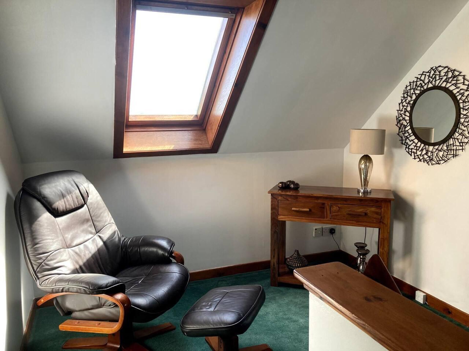Ein Zimmer mit Dachfenster, einem Ledersessel, einem Holztisch, einem Spiegel und einer Lampe.