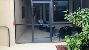 A covered patio area with a sliding glass door leading to an interior space. There are two black chairs and a small table, and a potted plant is visible in the foreground.