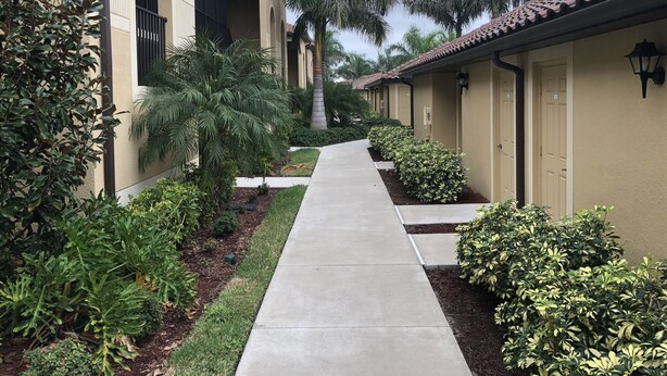 A pathway leading to a row of beige buildings with arched windows and a tiled roof, surrounded by well-maintained landscaping and palm trees.