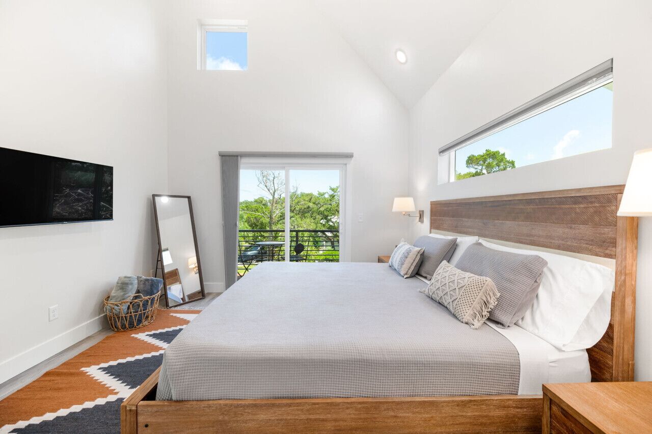 A bedroom with a large bed, a TV mounted on the wall, a window, and a balcony with a view of greenery.