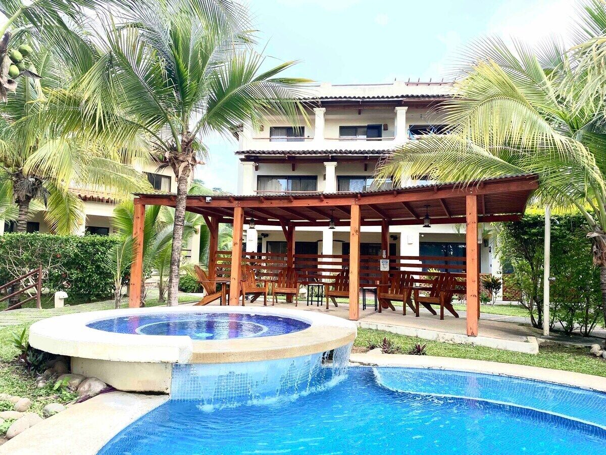 Piscina con jacuzzi, rodeada de palmeras y una pérgola de madera con sillas.