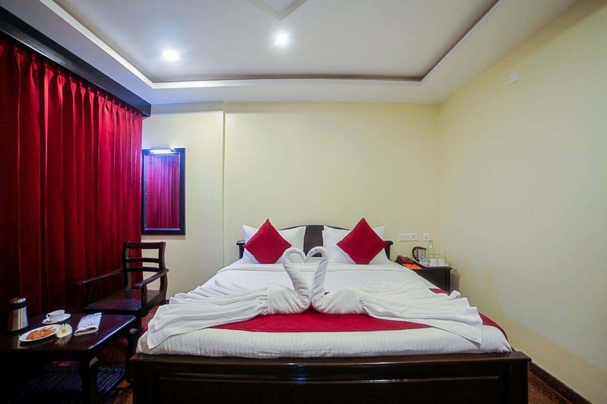 A hotel room with a neatly made bed, red curtains, a wooden desk, and a nightstand with a lamp.