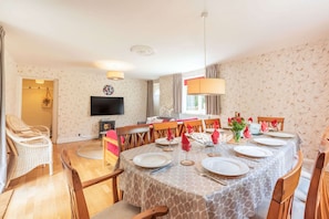 Un coin repas avec une table ronde dressée pour six personnes, entourée de chaises en bois. Un téléviseur est fixé au mur, et il y a une fenêtre avec des rideaux.