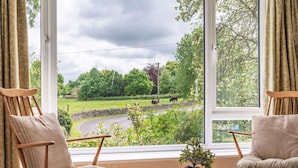 Ein Zimmer mit zwei Stühlen vor einem großen Fenster mit Blick auf eine ländliche Landschaft, Kühen und einer kurvigen Straße.