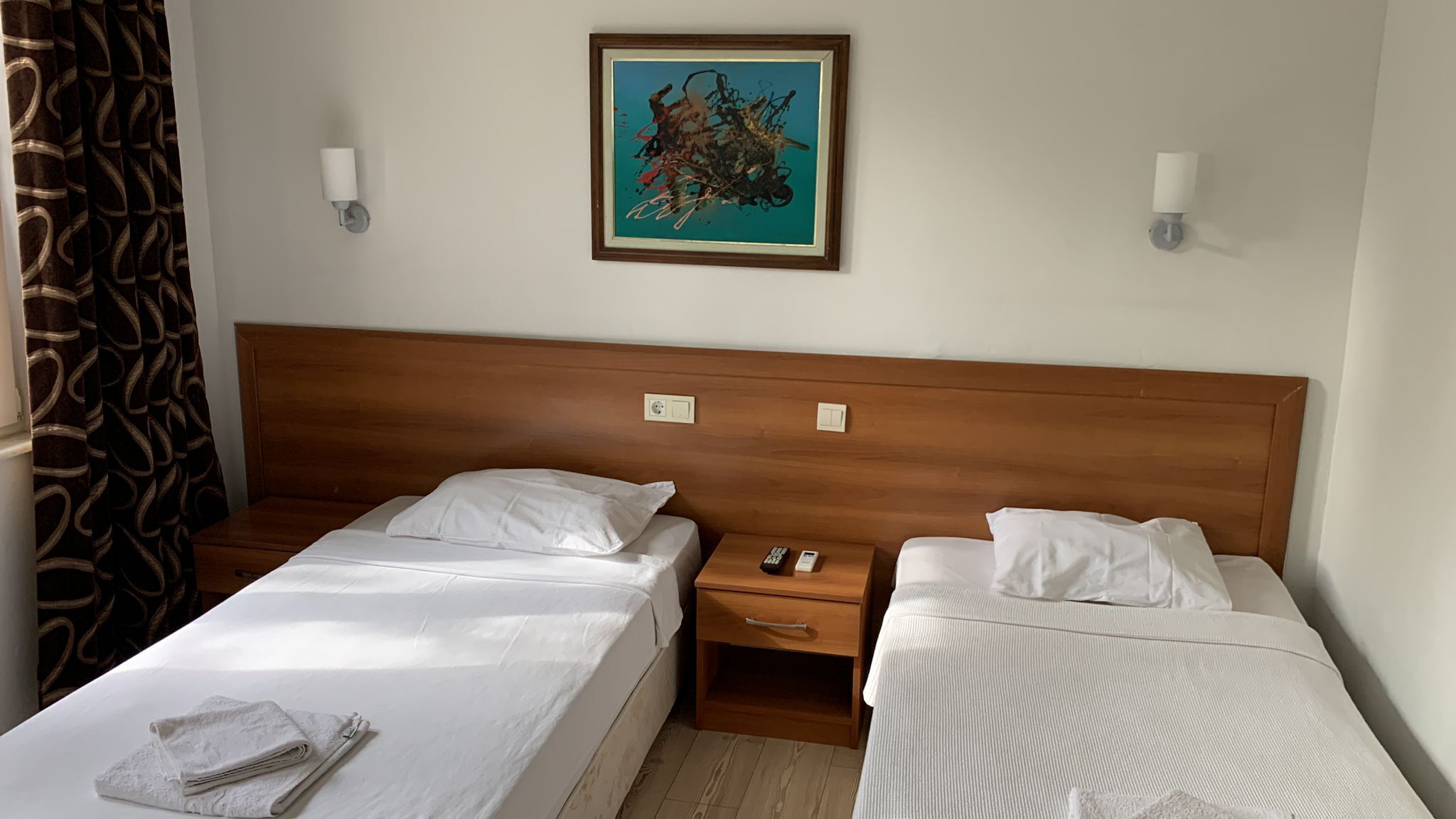 A hotel room with two beds, a wooden headboard, a painting on the wall, and a curtain with a leaf pattern.