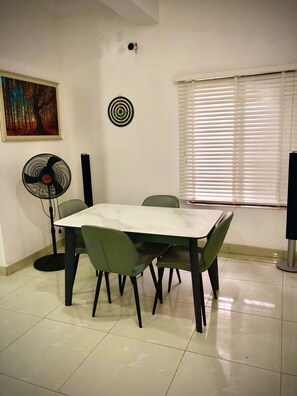 A dining area with a table and chairs, a standing fan, a dartboard, and a window with blinds. - Beautiful furnished 2 bedroom house in Abuja, FCT, Nigeria - Six Star (Abuja)