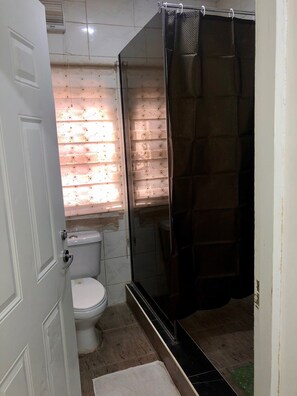A small bathroom with a toilet, a shower with a dark curtain, and a window with patterned blinds. - Beautiful furnished 2 bedroom house in Abuja, FCT, Nigeria - Six Star (Abuja)
