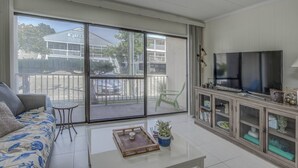 A living room with a sofa, a coffee table, a TV, and a sliding glass door leading to a balcony. - 15% off 5 nights or more (Ocean City)