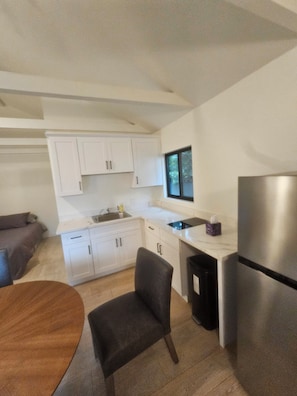 A compact kitchen with white cabinets, a stainless steel refrigerator, a small window, and a dining area with a wooden table and a black chair. - Westside Living! Contemporary Venice Beach Studio Suite (Los Angeles)