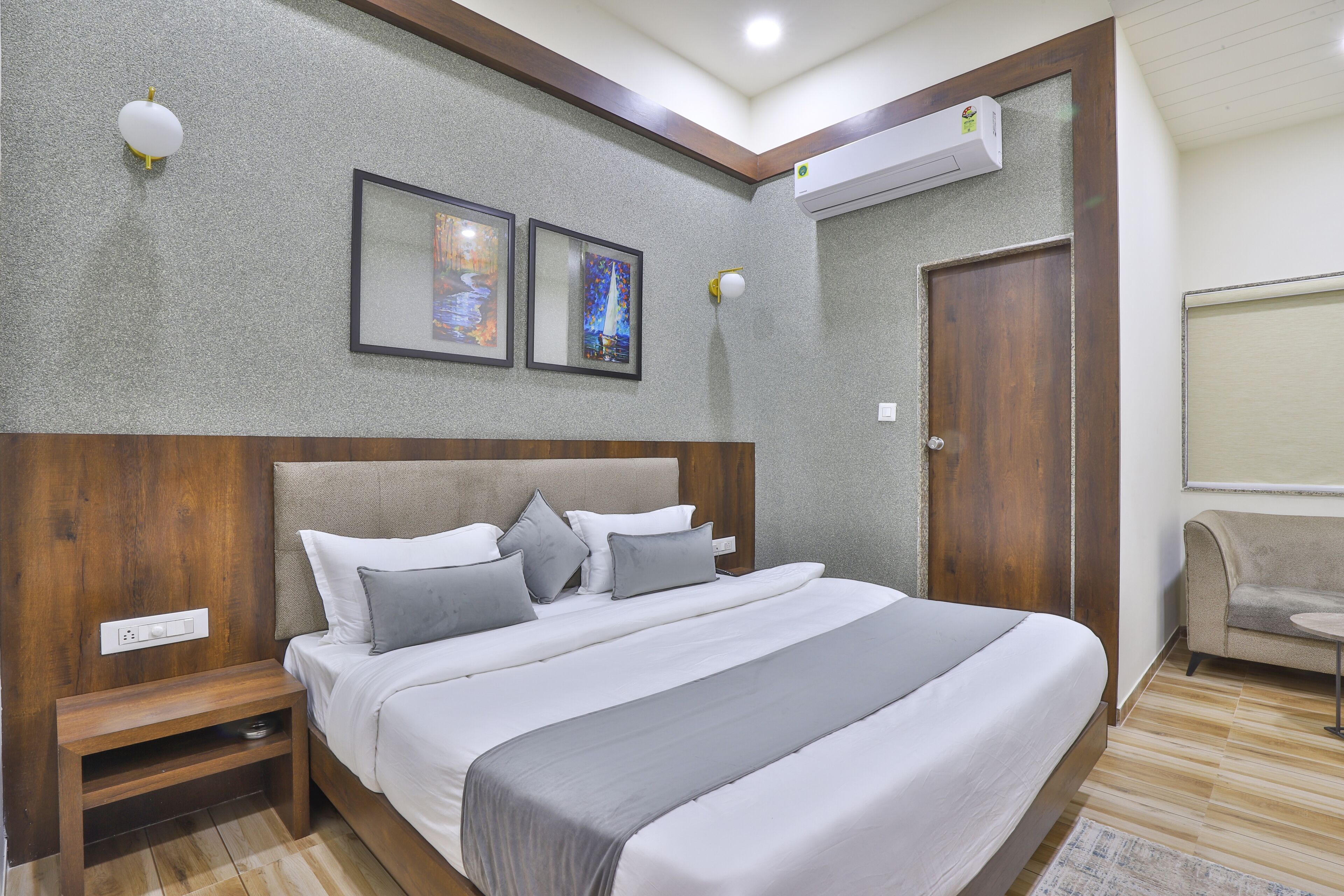 A modern bedroom with a large bed, a wooden nightstand, a wall-mounted air conditioner, and two framed artworks.