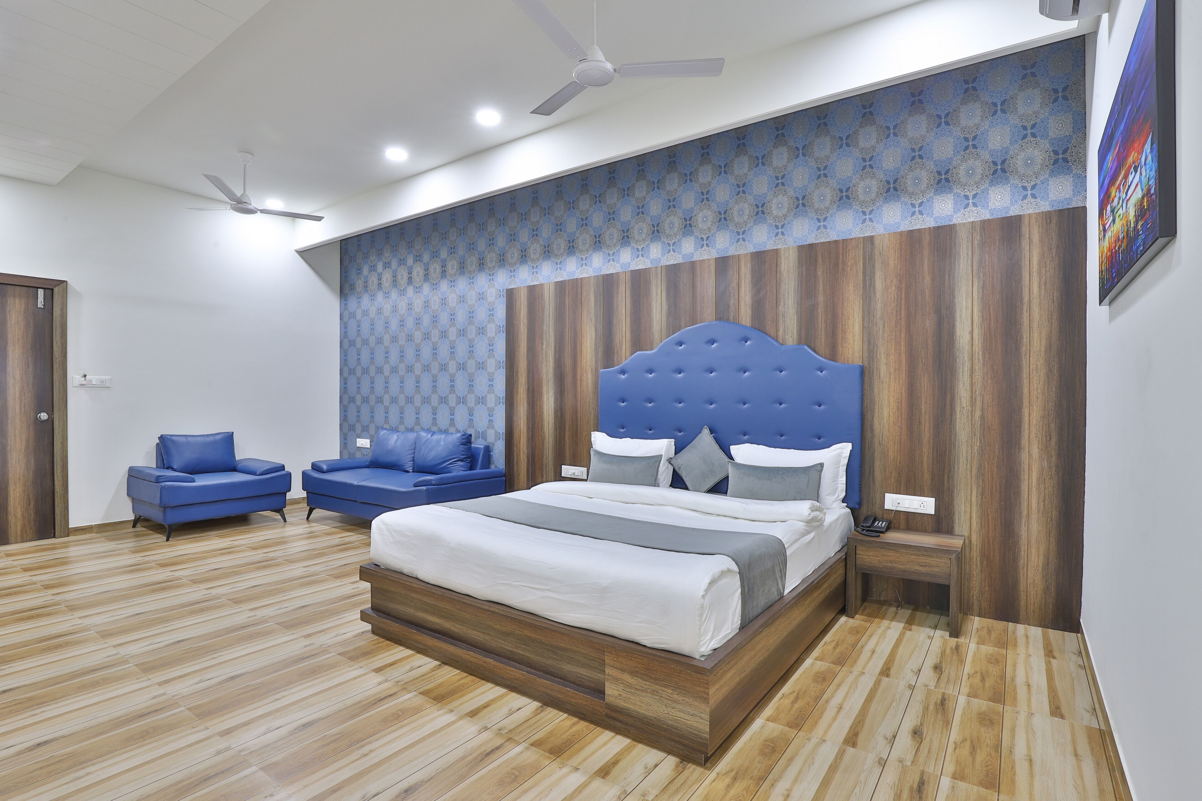 A modern bedroom with a large bed, two blue sofas, a wooden headboard, and a patterned wall.