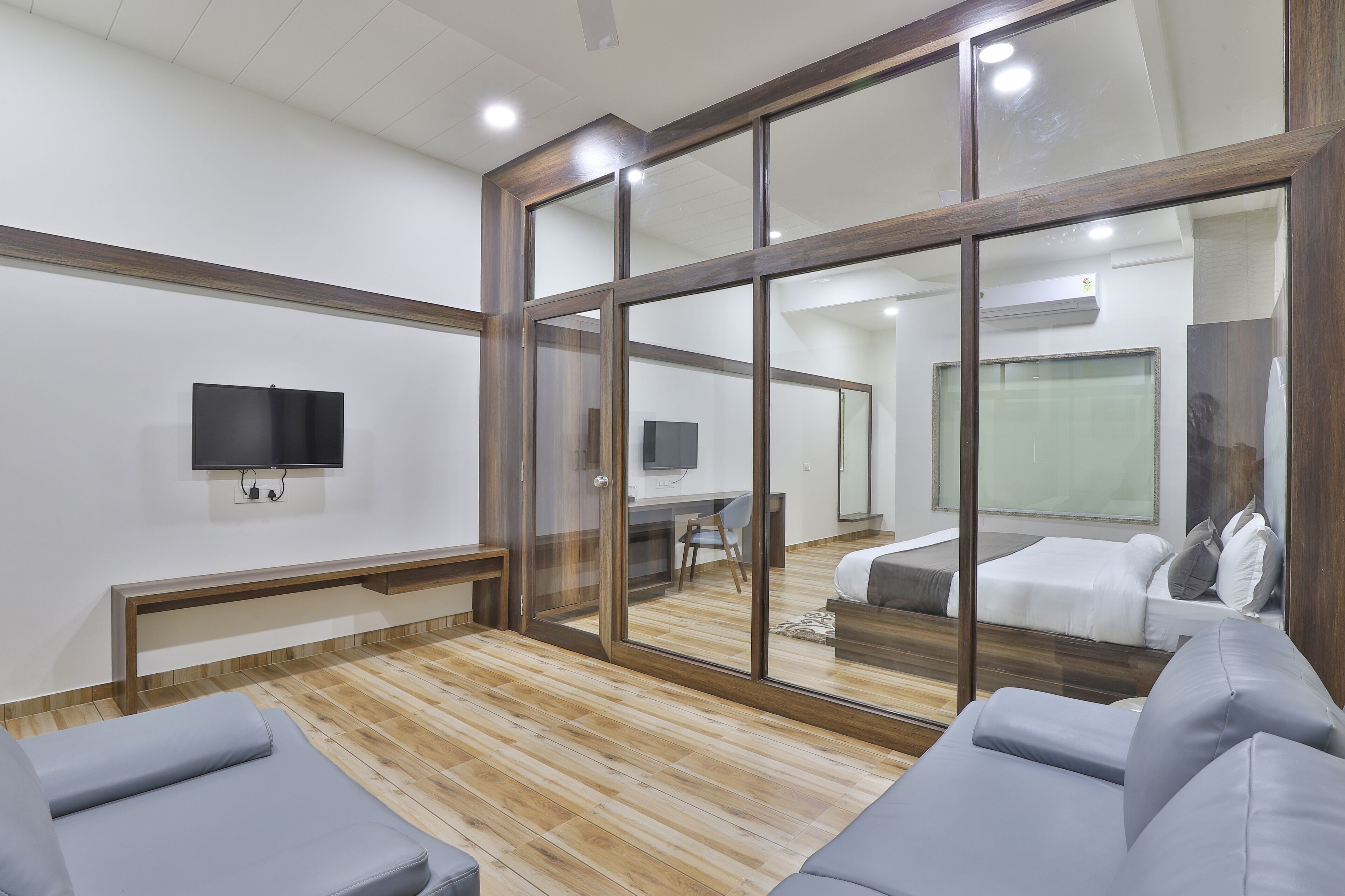 A modern hotel room with a large sliding glass door, a flat-screen TV mounted on the wall, a wooden floor, and a grey sofa.