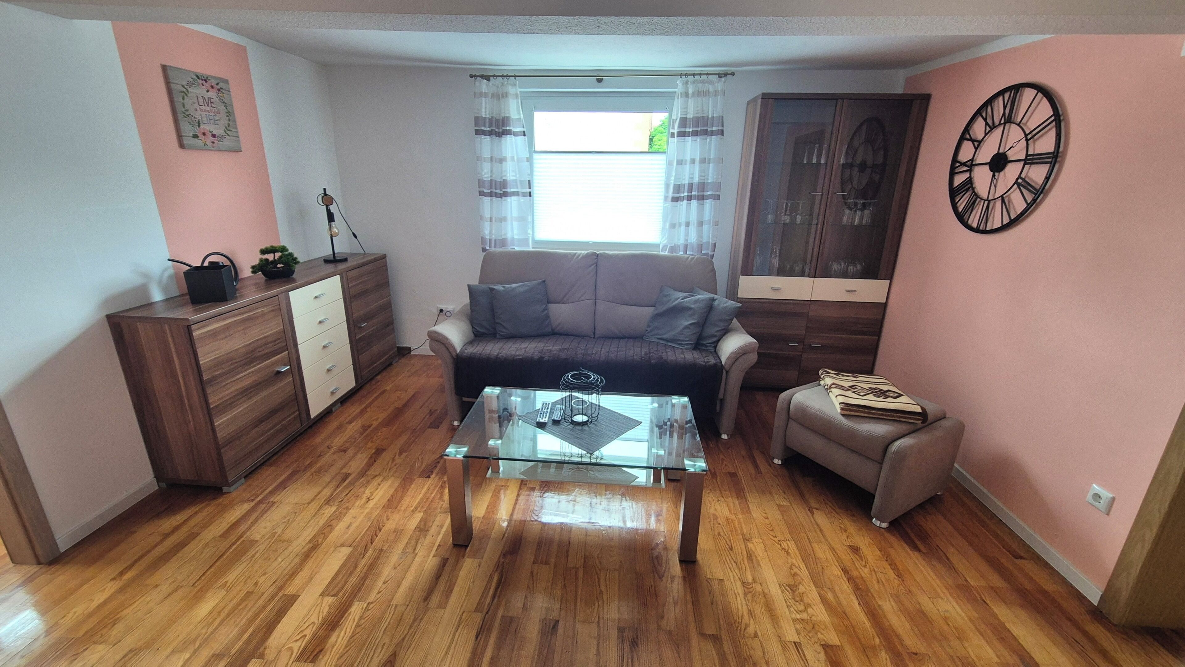 A living room with a sofa, a glass coffee table, a wooden cabinet, a small table with a lamp, and a clock on the wall.