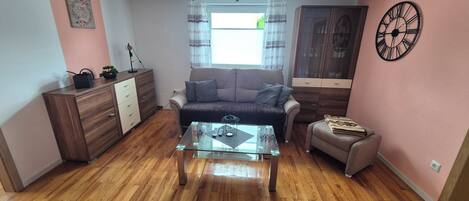A living room with a sofa, a glass coffee table, a wooden cabinet, a small table with a lamp, and a clock on the wall.
