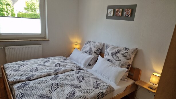 Een bed met een sprei met bladmotief, twee bedlampjes en een raam met uitzicht op groen.