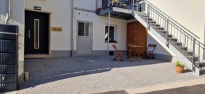 Un bâtiment avec une porte noire, un escalier en bois et un petit espace extérieur aménagé avec deux chaises et une table.