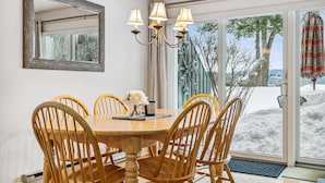 Un coin repas avec une table ronde en bois, six chaises en bois, un lustre, un miroir et une vue sur un paysage enneigé par de grandes portes vitrées.