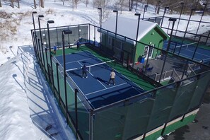 Un court de tennis avec un filet, entouré d’une clôture verte, et un petit bâtiment avec un toit vert.