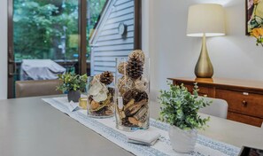 A dining table with two glass vases containing natural elements, a potted plant, and a lamp on a wooden side table.