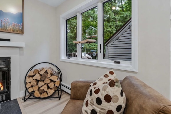 A living room with a fireplace, a window overlooking a deck, and a stack of firewood.