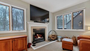A living room with a fireplace, a TV, and a large window. - Fireplace Condo, Pet-Friendly, Newton Village (Quechee)