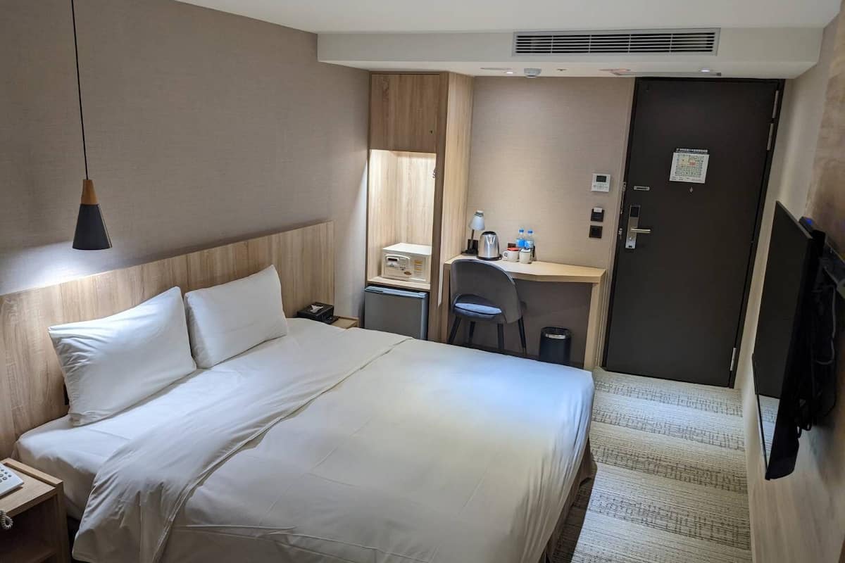 A hotel room with a large bed, a desk with a chair, a desk lamp, a telephone, and a door.