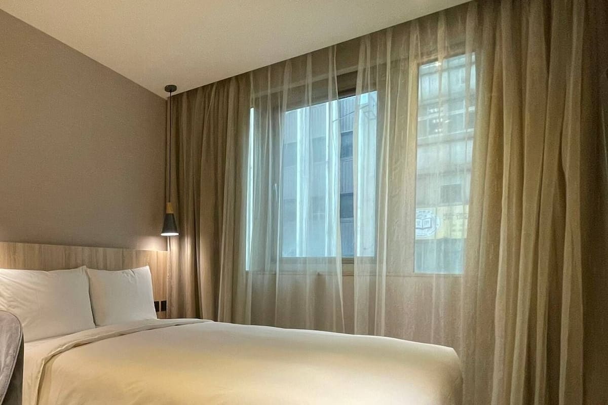 A hotel room with a bed, a window with sheer curtains, and a wall-mounted lamp.