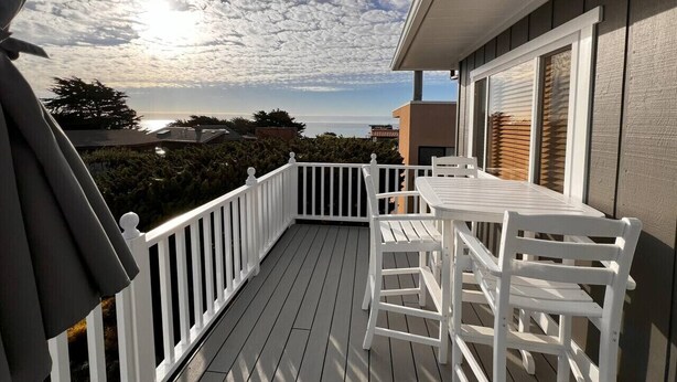 A wooden deck with white railings and a table with chairs, overlooking a residential area and a body of water.