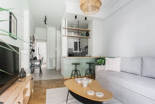 A modern living room with a grey sofa, a wooden coffee table, and a TV on a wooden stand. There is a kitchen area with a counter and stools. - Dazzling 1BR Apartment in Koukaki (Athens)