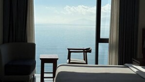 Une pièce comprenant un lit, une chaise et une petite table, avec une grande fenêtre donnant sur la mer et sur une montagne lointaine.
