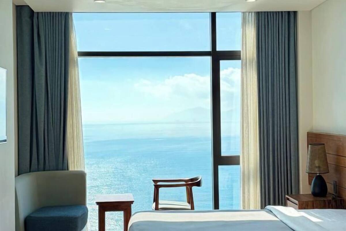 Une chambre d’hôtel avec une grande fenêtre, un lit, une chaise, une petite table et une lampe.