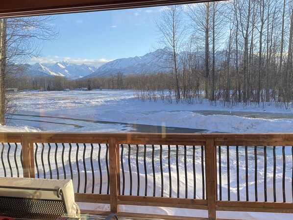 A view from a wooden deck overlooking a snow-covered landscape with a river and mountains in the background.