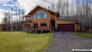A large wooden cabin with a prominent front porch, a garage, and a clear view of the surrounding forest. - Lazy Mountain View- Spacious log home with mountain views, just north of Palmer (Palmer)
