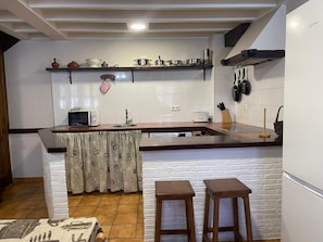 Eine Küche mit weiß gefliestem Spritzschutz, einer Holzarbeitsplatte, einer Mikrowelle, einem Toaster und einem Spülbecken. Zwei hölzerne Hocker und ein kleiner Tisch mit gemustertem Tischläufer sind ebenfalls vorhanden.
