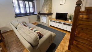 Una sala de estar con un sofá, un televisor sobre un mueble de madera, una chimenea de piedra, una ventana con cortinas y una escalera.