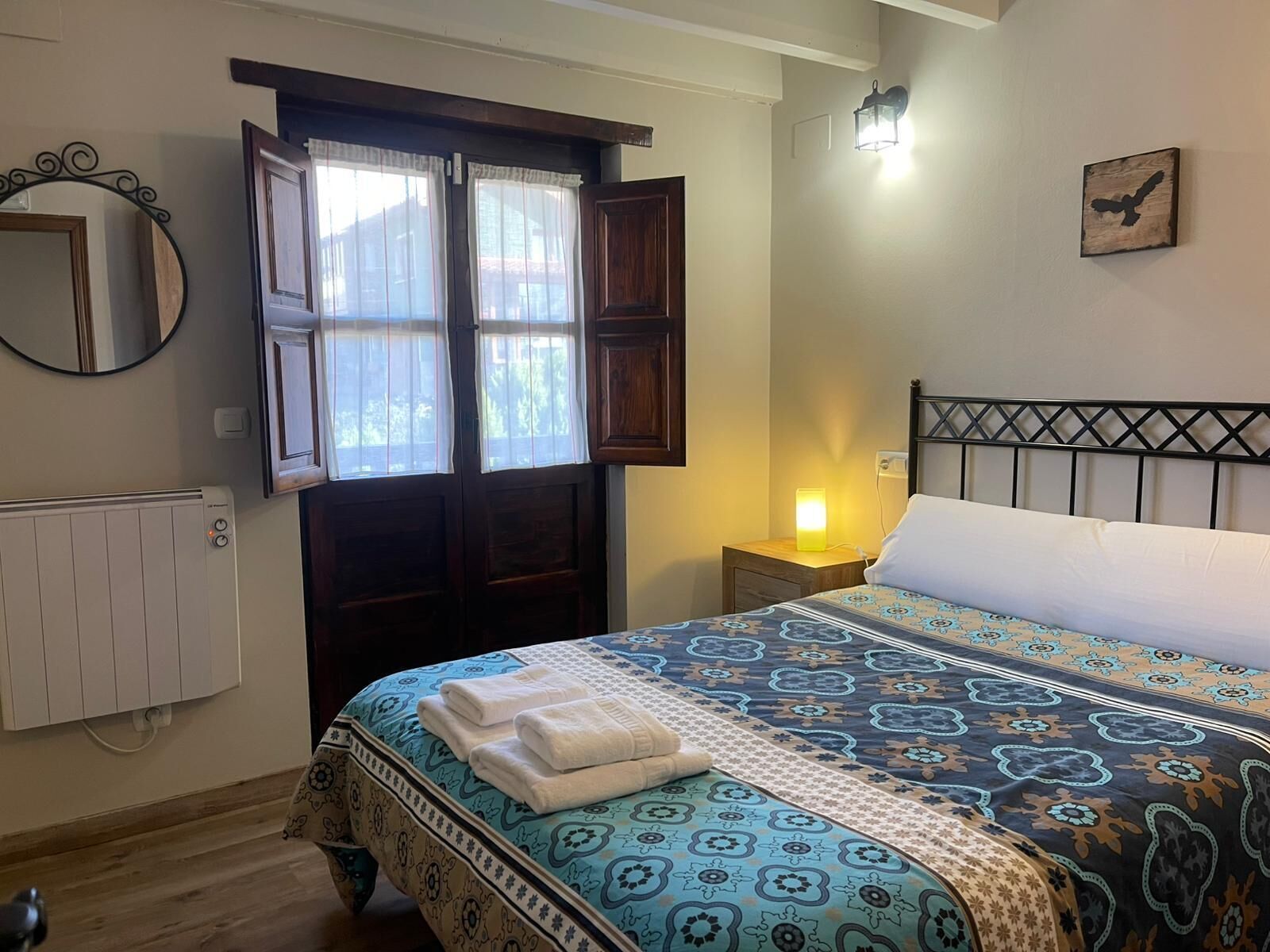 A bedroom with a bed, a nightstand, a wall-mounted lamp, a window with curtains, a radiator, and a mirror.