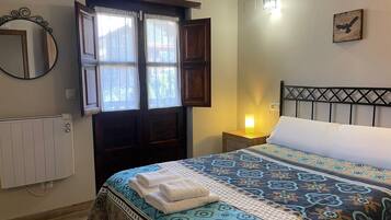 A bedroom with a bed, a nightstand, a wall-mounted lamp, a window with curtains, a radiator, and a mirror.