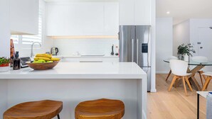 A modern kitchen with white cabinetry, a stainless steel refrigerator, a glass dining table with white chairs, and wooden stools. - Top Floor Apartment Close To Bondi Beach (Bondi)
