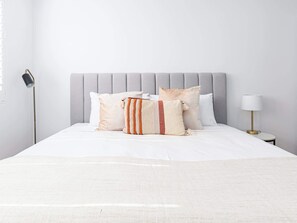 Un lit avec une tête de lit rembourrée de couleur grise, une literie blanche et un plaid à rayures.