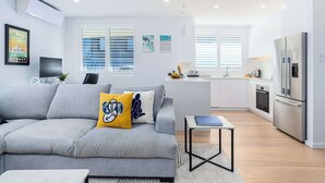 A modern living room with a grey sofa, a small table, and a kitchen area with white cabinets and a stainless steel refrigerator. - Top Floor Apartment Close To Bondi Beach (Bondi)